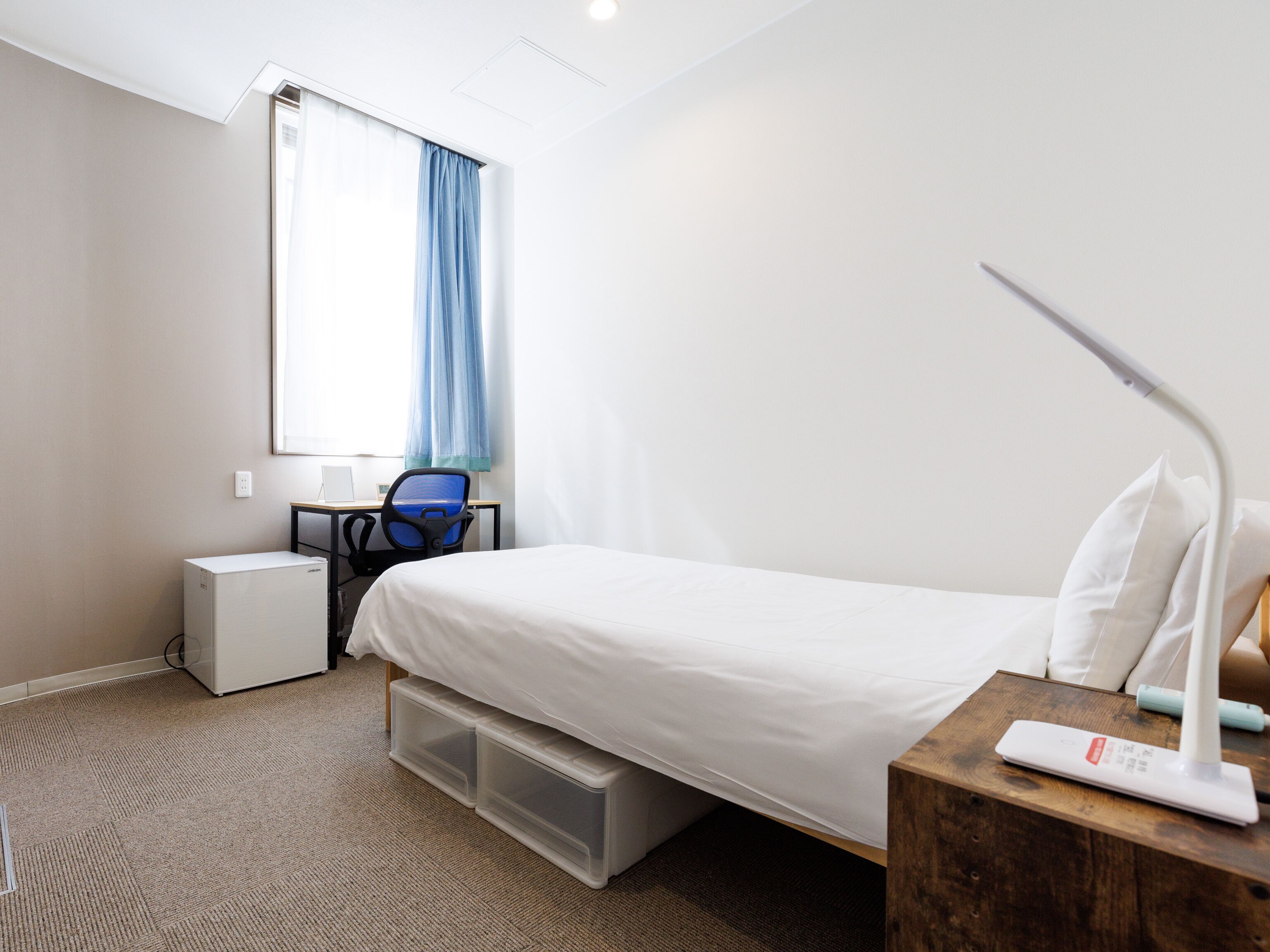 A hotel room with a bed, a desk with a chair, a window with curtains, a bedside table with a lamp, and storage boxes under the bed.