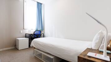 A hotel room with a bed, a desk with a chair, a window with curtains, a bedside table with a lamp, and storage boxes under the bed.