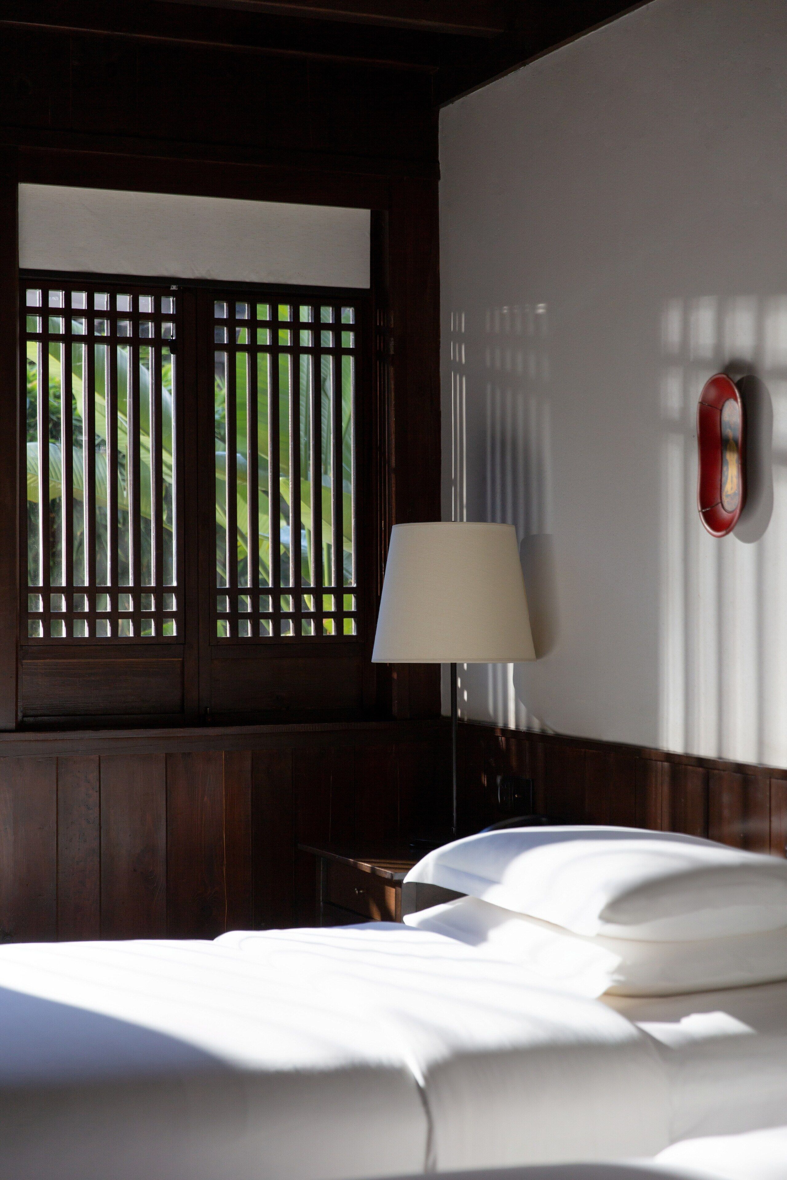 A hotel room with a bed, a nightstand, a lamp, and a window with a view of greenery.