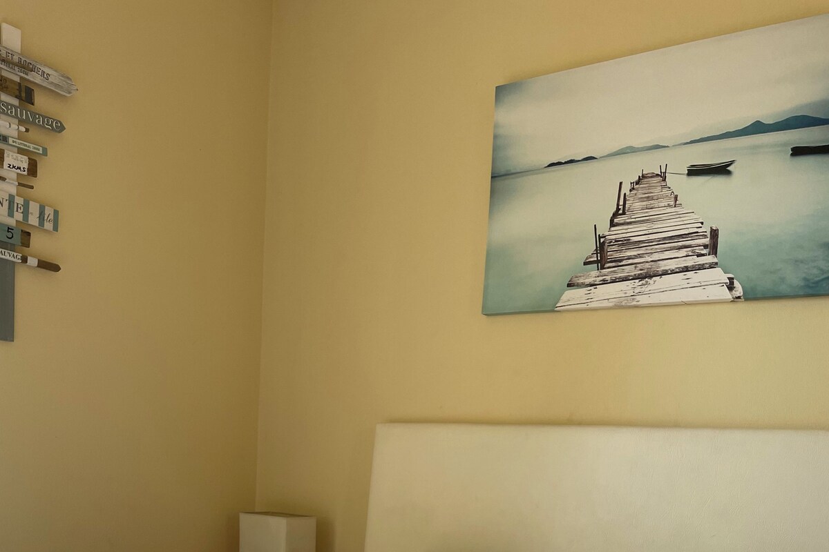 Habitación de hotel con cama, almohadas, un cuadro de un muelle y una toalla amarilla con texto bordado.