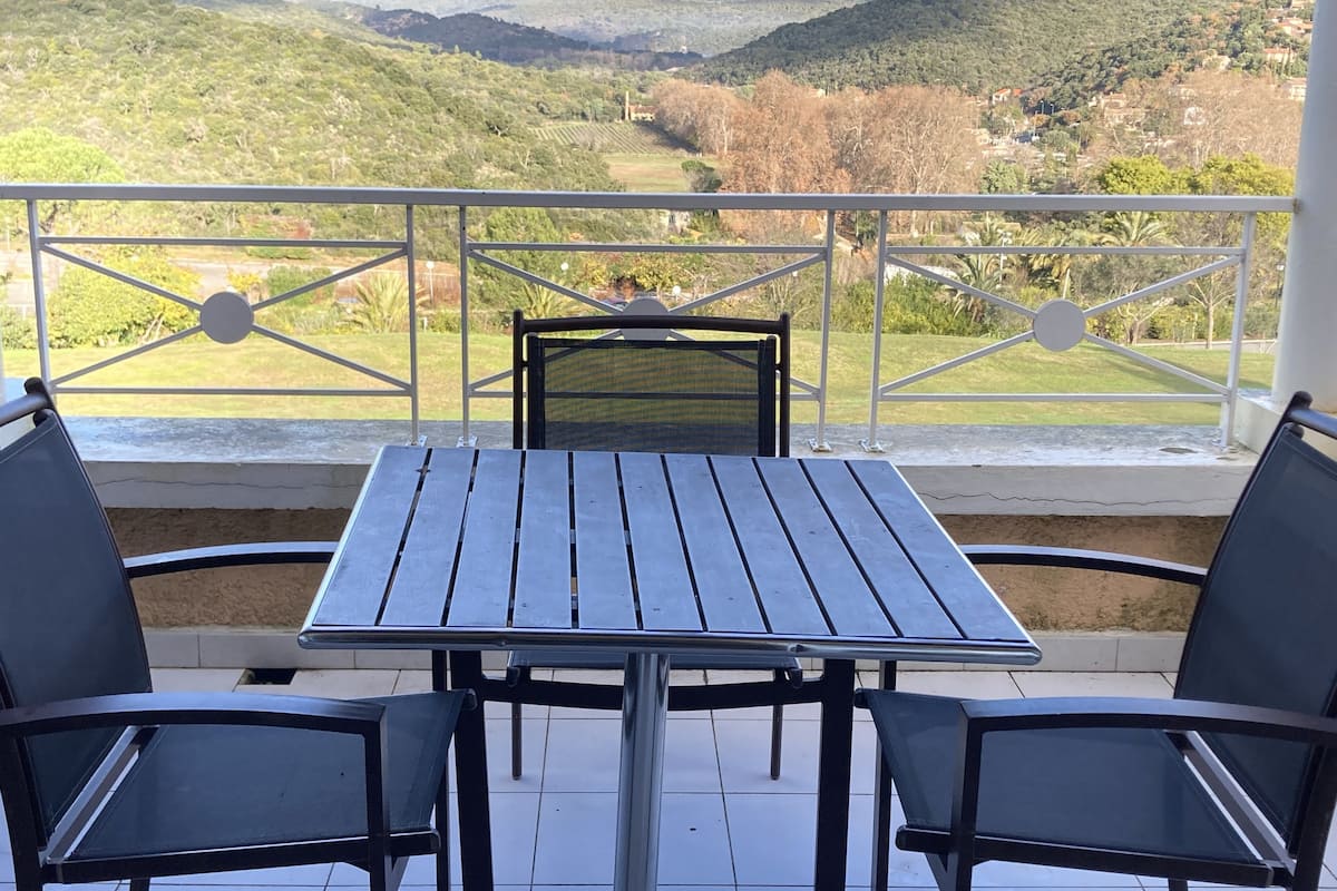 A balcony with a table and chairs, offering a view of a green landscape with hills.