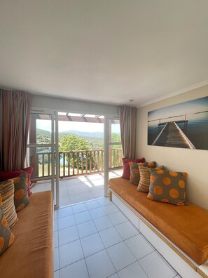 A balcony with a view of greenery and mountains, a sofa with patterned cushions, and a painting on the wall. - Residence Cap Esterel By Actisource  (Saint-Raphael)