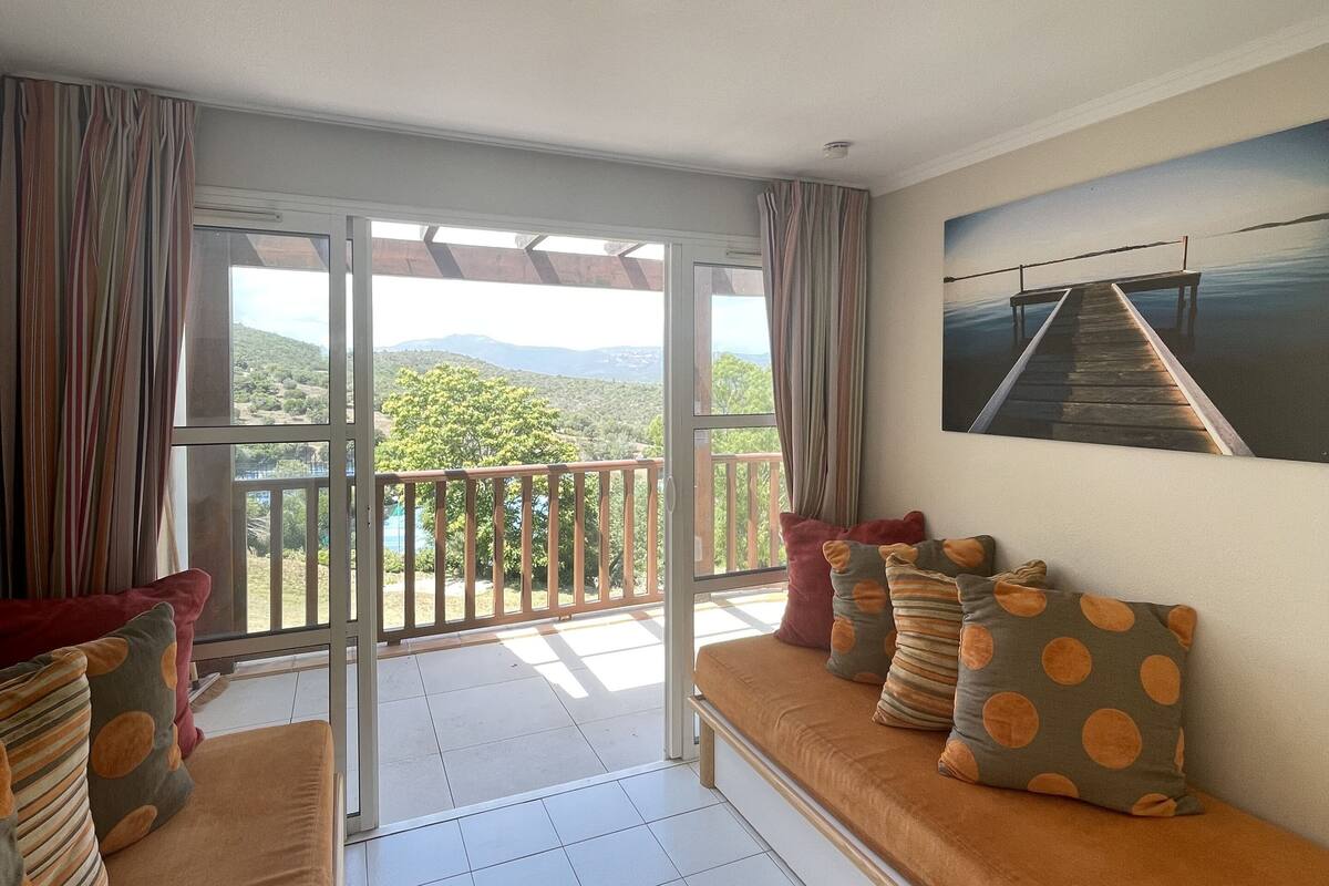 A balcony with a view of greenery and mountains, a sofa with patterned cushions, and a painting on the wall.