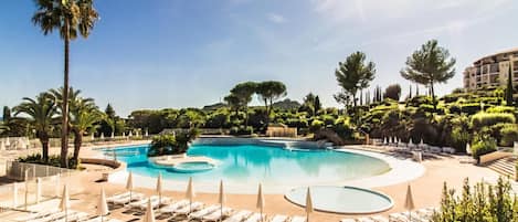 Une grande piscine incurvée, dotée de chaises longues et de parasols, et entourée de palmiers et d’autres plantes.