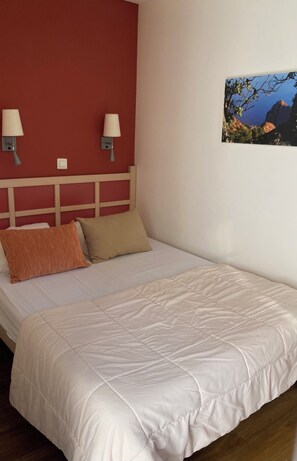 Una cama individual con edredón blanco, dos almohadas, cabecera de madera, dos lámparas fijadas a la pared y un cuadro enmarcado.