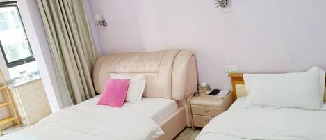 A hotel room with two beds, a window with curtains, an air conditioning unit, a bedside table with a lamp, and a wall-mounted light fixture.