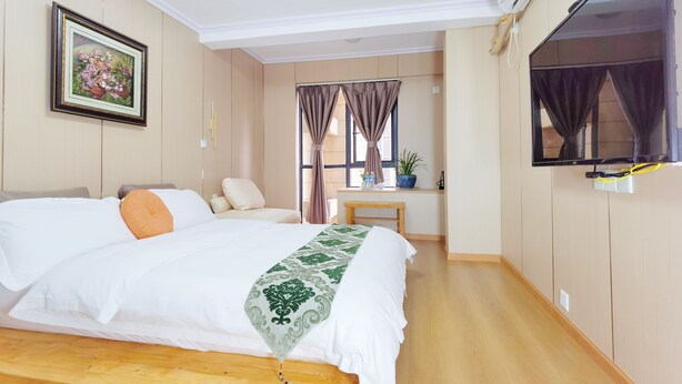 Habitación de hotel con una cama grande, un televisor montado en la pared, una ventana con cortinas y una mesita con una planta.