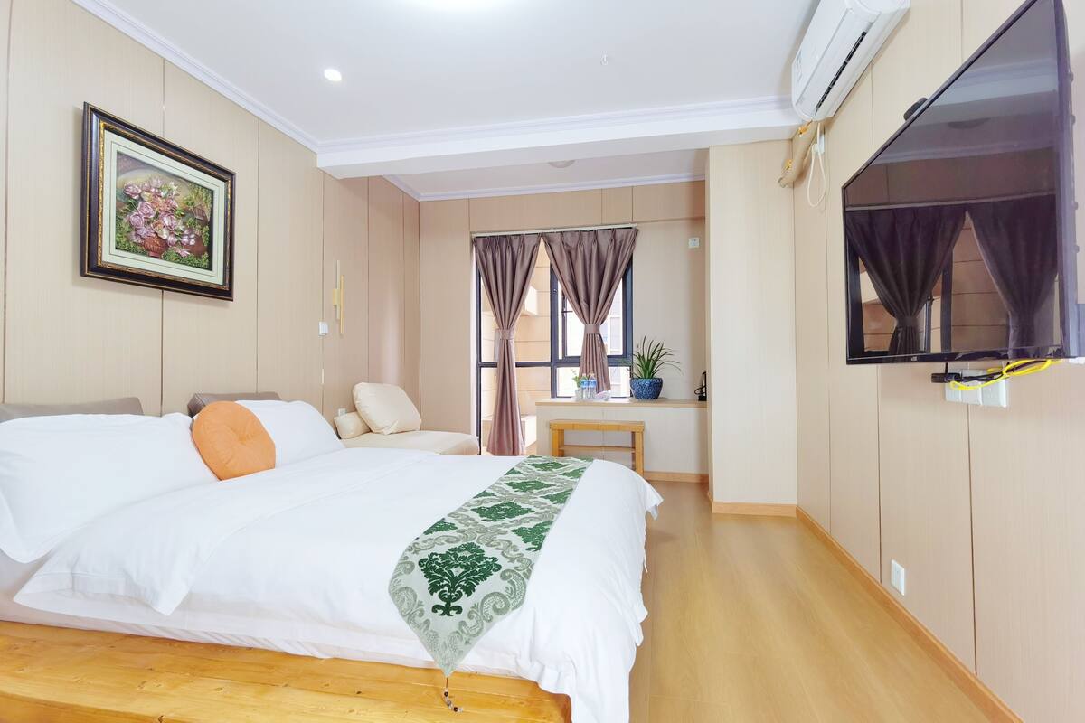 Habitación de hotel con una cama grande, un televisor montado en la pared, una ventana con cortinas y una mesita con una planta.