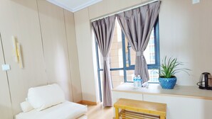 Una habitación ordenada con una cama, una mesita, un banco, un ventanal con cortinas y una planta en maceta.
