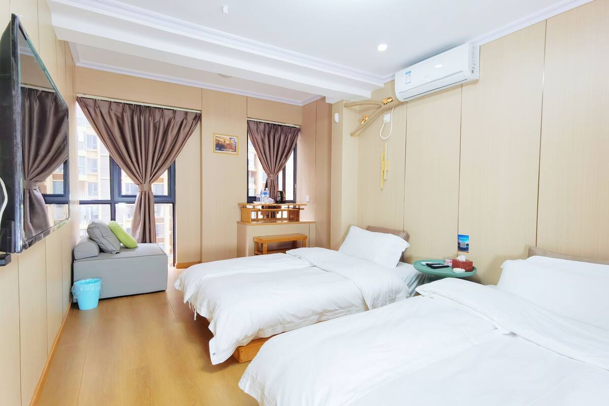 Habitación de hotel con dos camas, un pequeño rincón de descanso, un banco de madera, una mesita de noche y una ventana con cortinas.