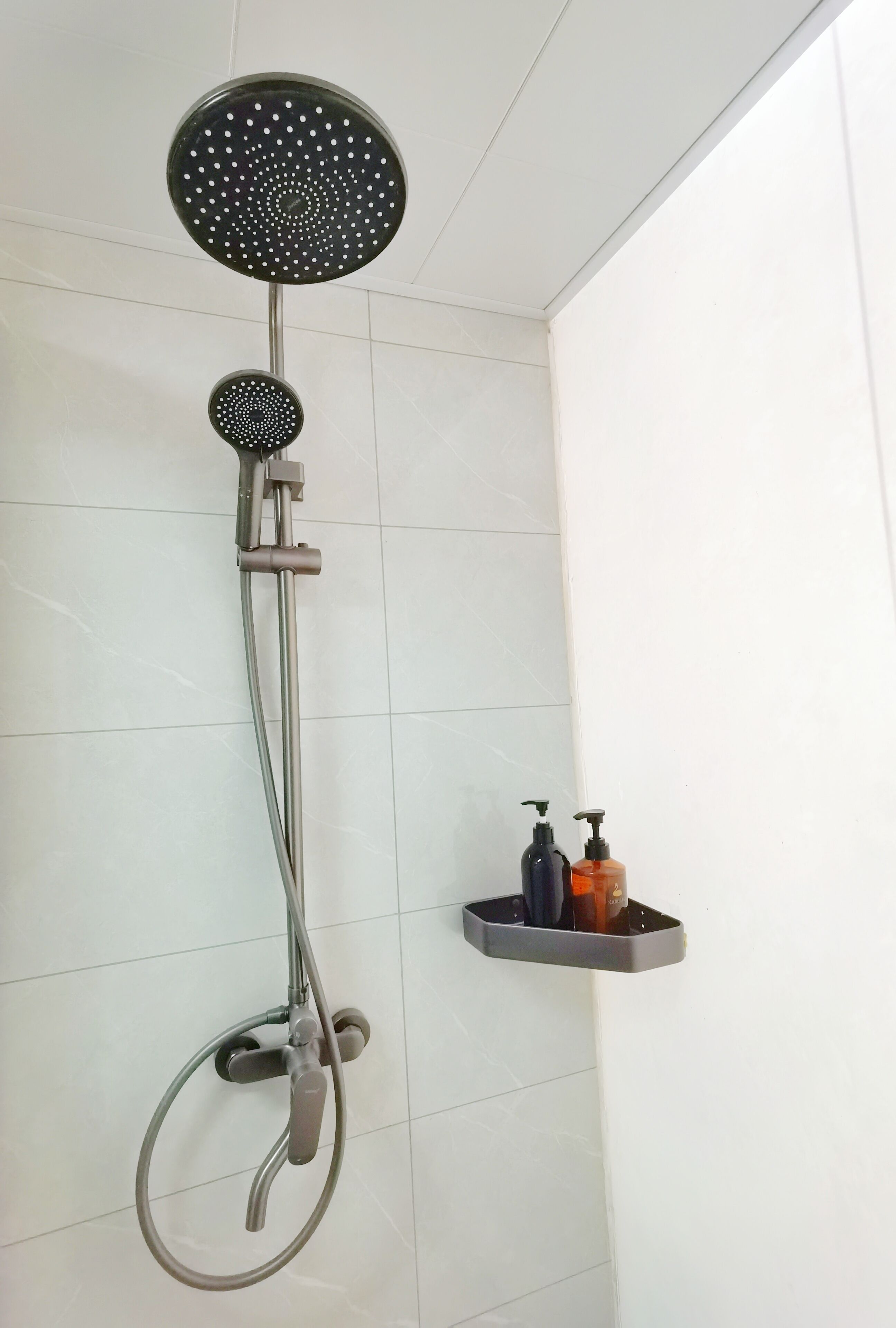 A shower with a handheld showerhead and a wall-mounted faucet. There is a shelf with two bottles of soap.