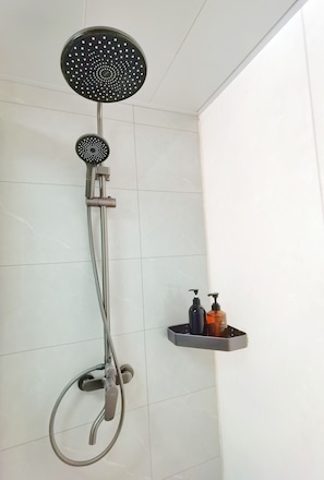 A shower with a handheld showerhead and a wall-mounted faucet. There is a shelf with two bottles of soap.