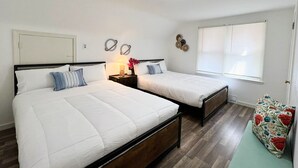 A bedroom with two beds, a nightstand with a lamp, a wall clock, and a window with blinds.