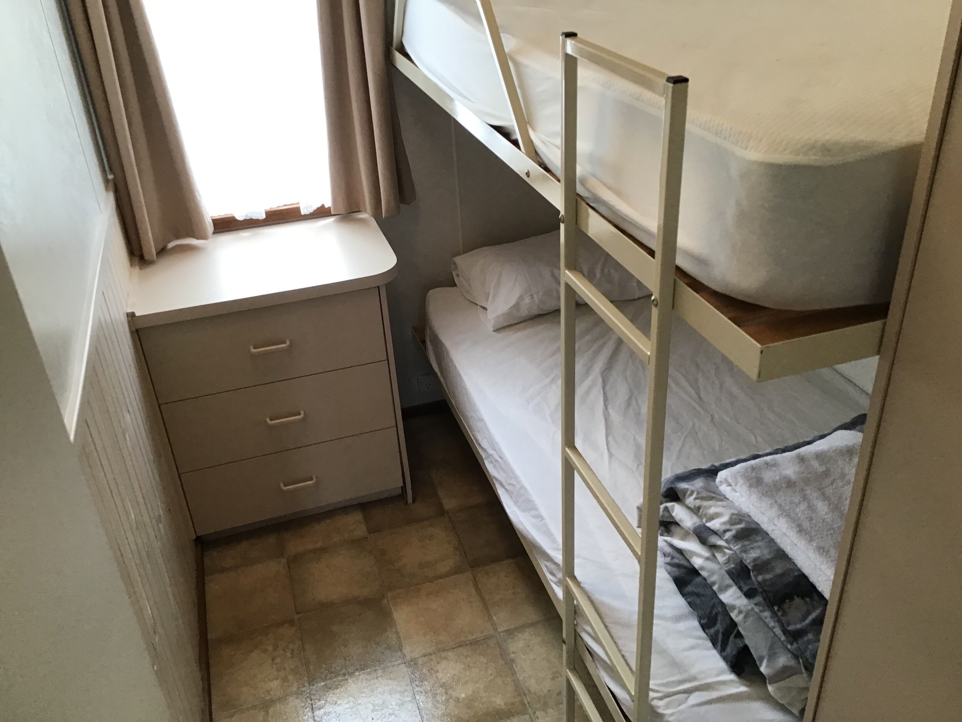 Une petite pièce, de taille modeste, comprenant un lit superposé, une table de chevet avec des tiroirs et une fenêtre avec des rideaux.