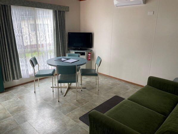 Une petite pièce bien éclairée, avec une table ronde, quatre chaises, un canapé vert, un téléviseur à écran plat et un extincteur.