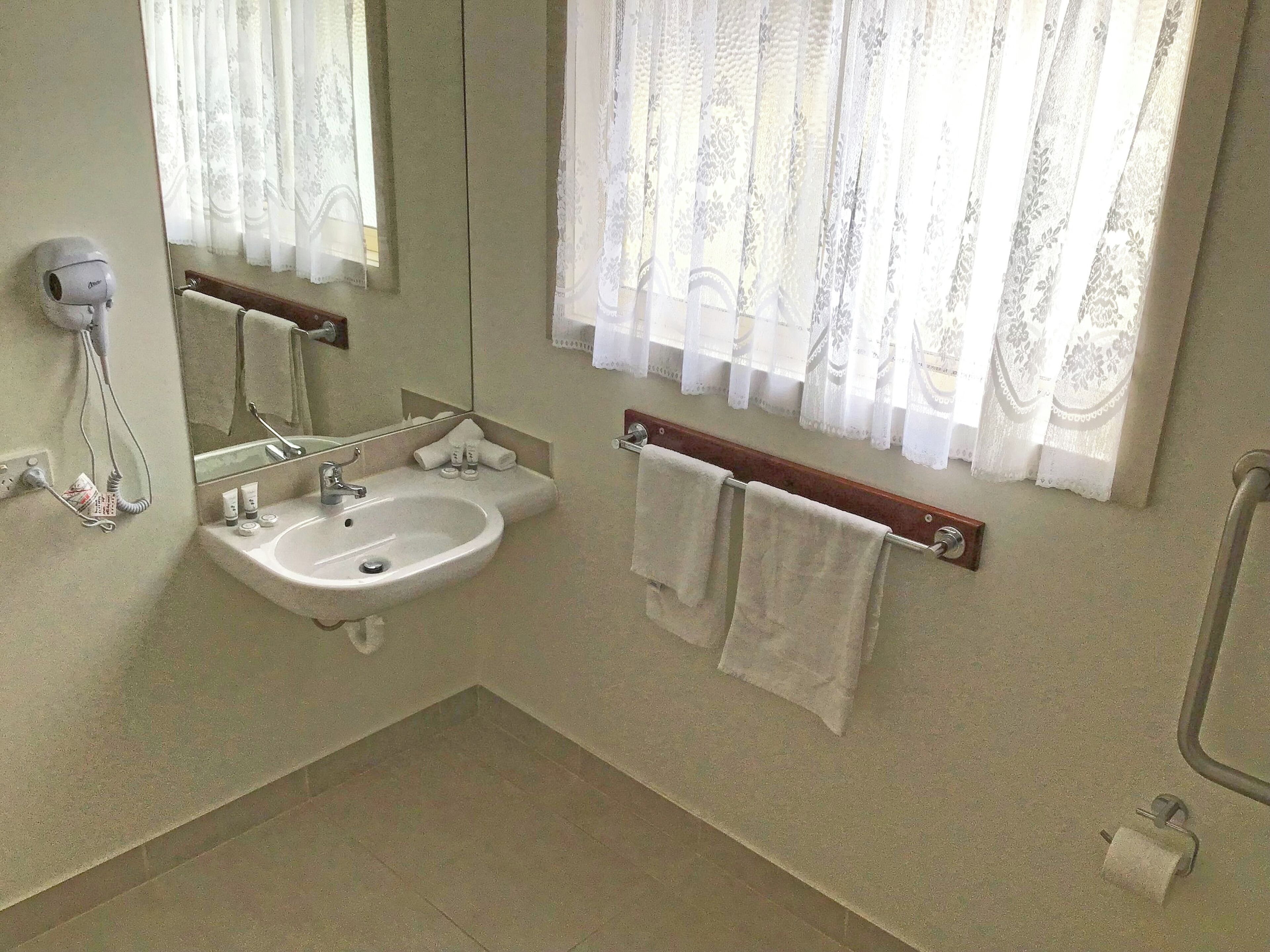 Un baño con lavamanos, espejo, toallero y una ventana con cortinas de encaje.
