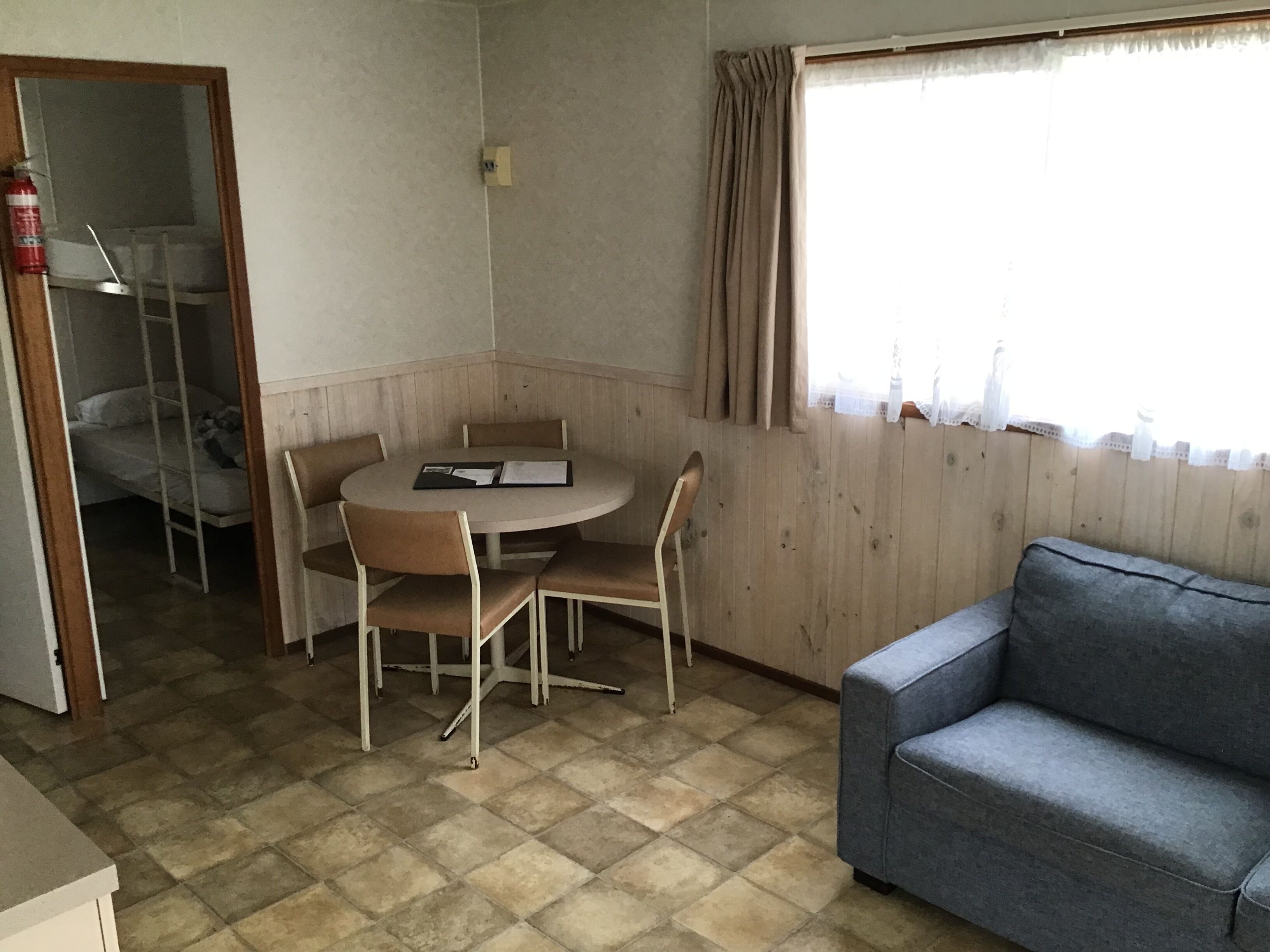 A room with a round table, four chairs, a sofa, a bunk bed, and a window with curtains.