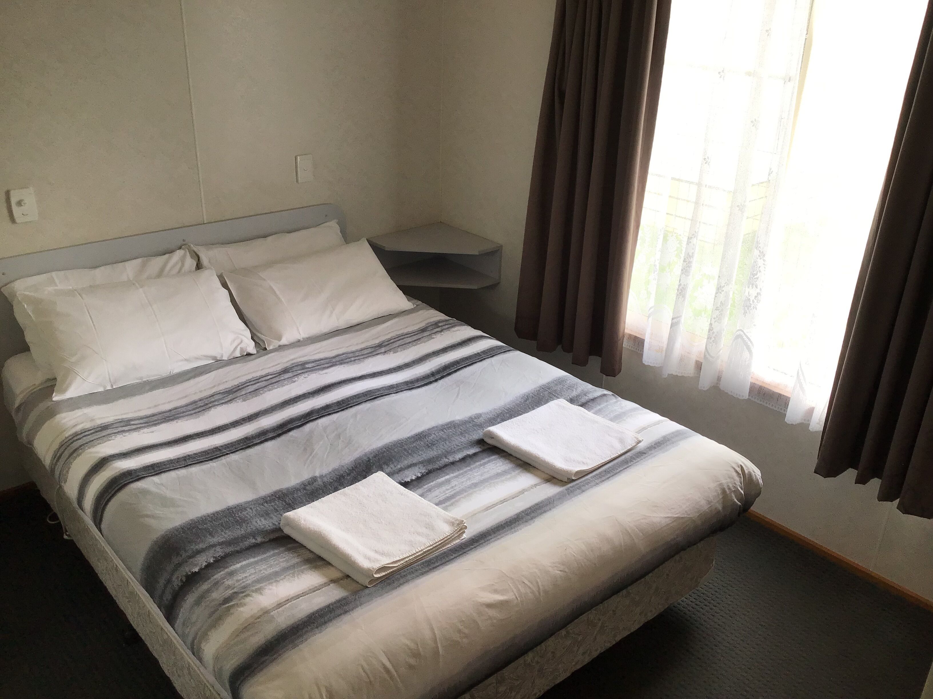 A neatly made bed with striped bedding and two folded towels, a bedside table, and a window with curtains.