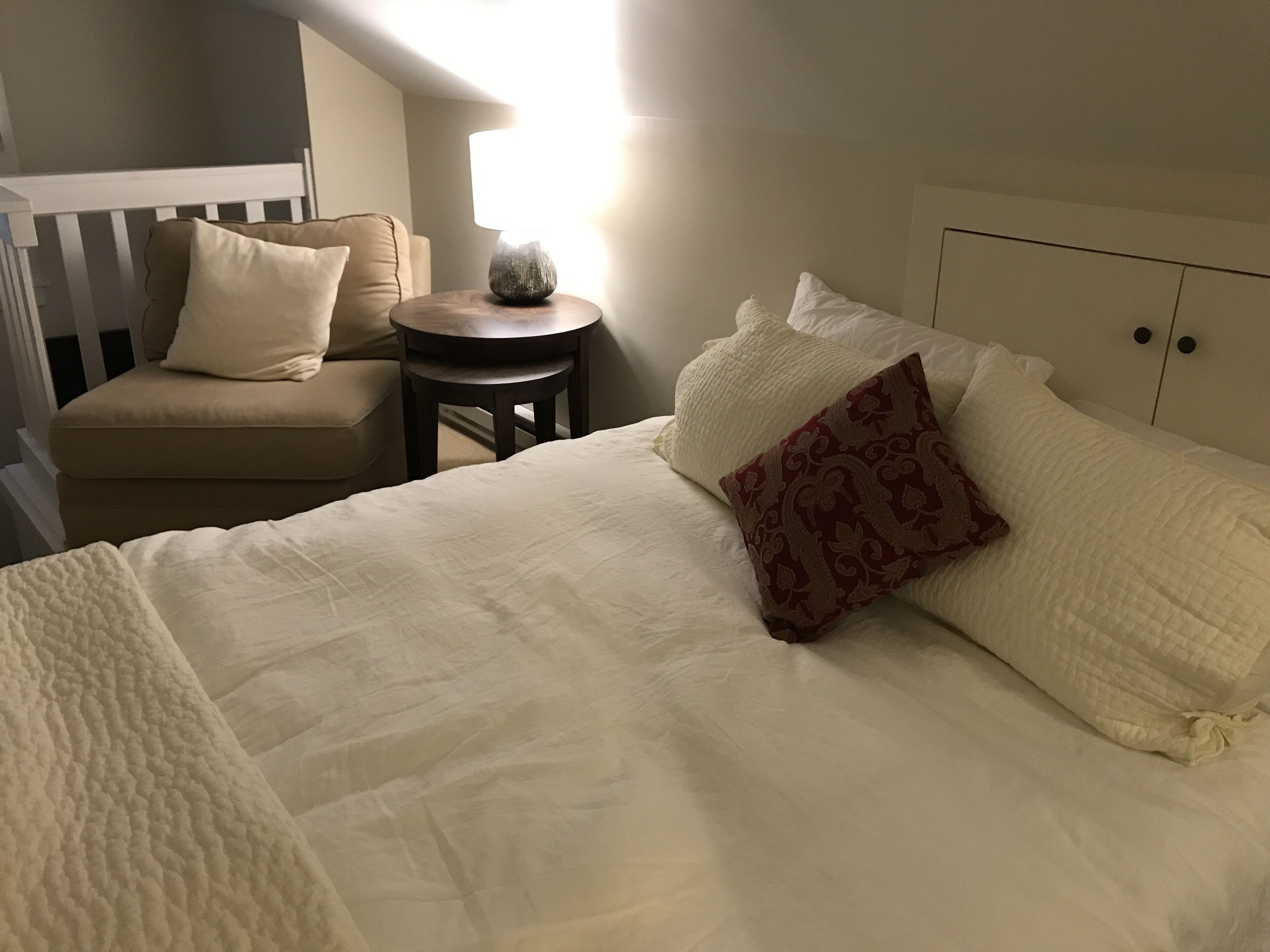 A neatly made bed with a patterned pillow, a small round table, and a chair in a room with a white headboard and a white railing.