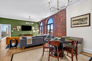 A modern living room with a dining area, green walls, a brick accent wall, and a large window.