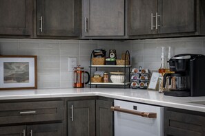 Une cuisine avec des meubles de couleur sombre, un plan de travail blanc, une machine à café, une cafetière et une étagère contenant divers objets.