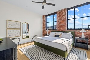 A modern bedroom with a large bed, a ceiling fan, a brick wall, and large windows.