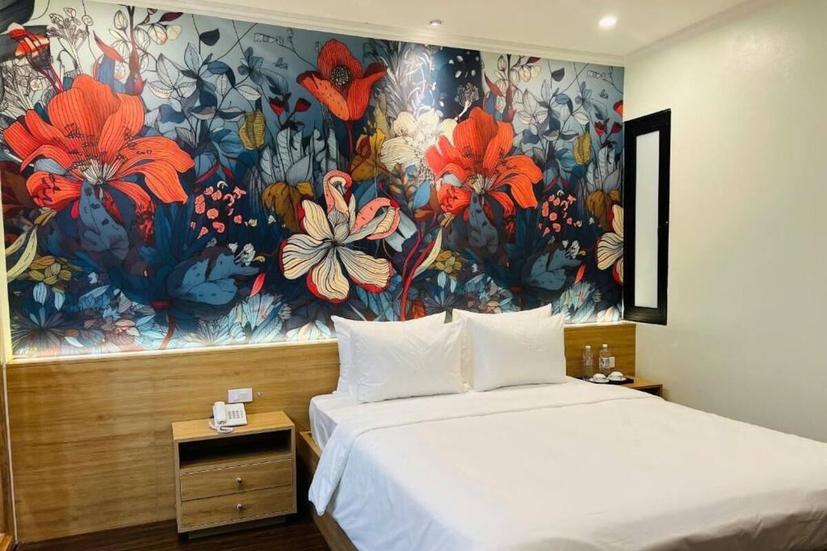 Ein Hotelzimmer mit einer großen floralen Wandmalerei, einem hölzernen Kopfteil, einem Bett mit weißen Leinen, einem Nachttisch mit einem Telefon und einem Beistelltisch mit einer Lampe.