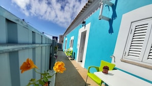 Un estrecho pasillo exterior con una pared azul, una puerta blanca y una silla verde.