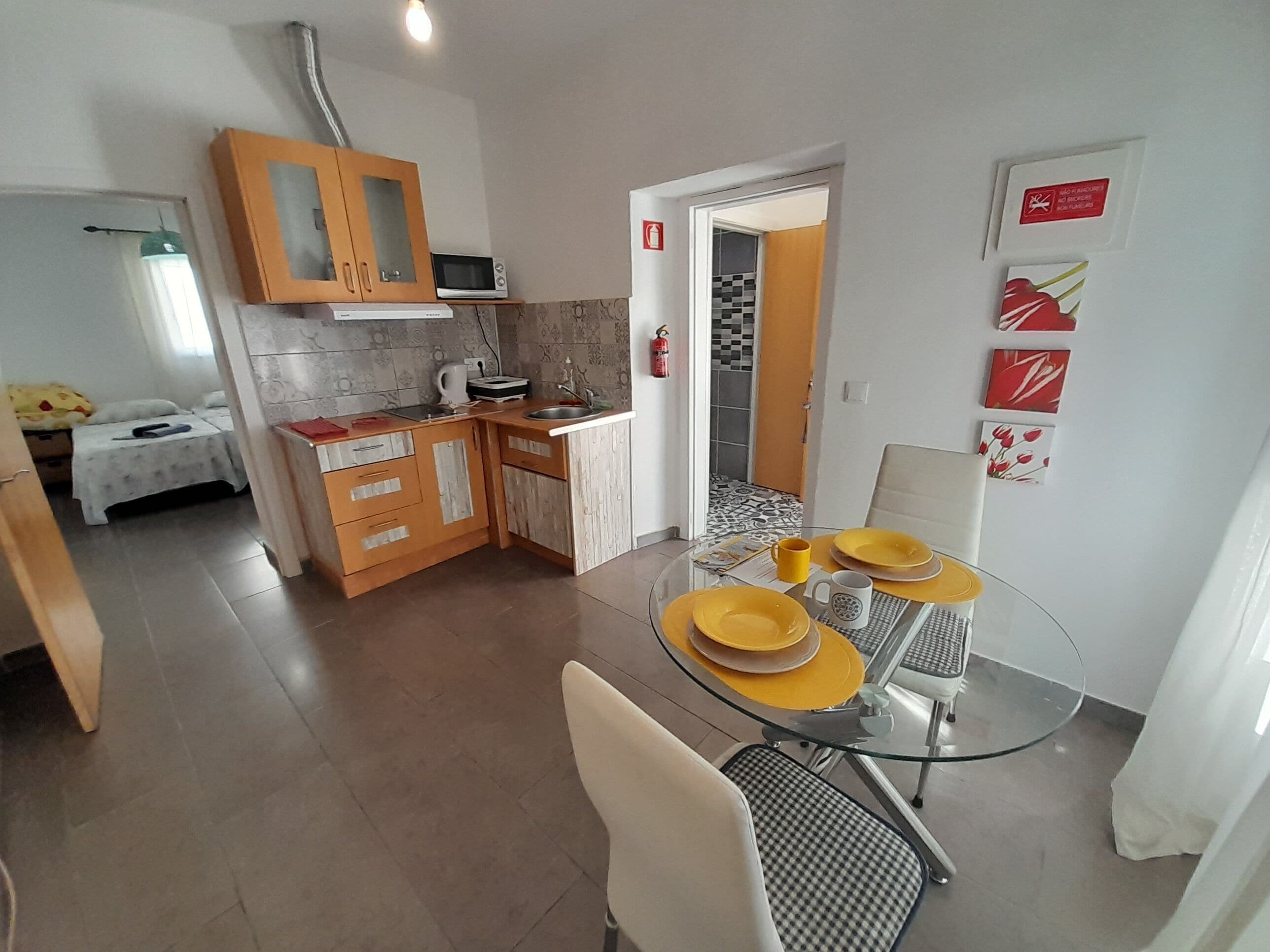 A compact kitchen with wooden cabinets, a microwave, and a sink. There's a dining area with a glass table and chairs, and a bed with a yellow blanket in the background.