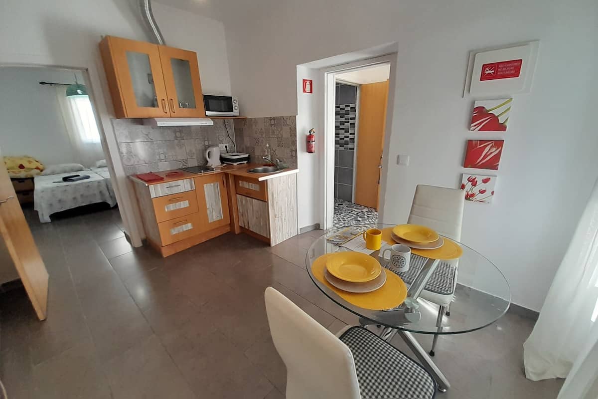 Une cuisine compacte avec des meubles en bois, un four à micro-ondes et un évier. On y trouve un coin repas avec une table et des chaises en verre, et un lit avec une couverture jaune en arrière-plan.