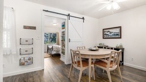 Et spiseområde med et rundt bord og stole, en ventilator i loftet og udsigt til et rum med et juletræ.