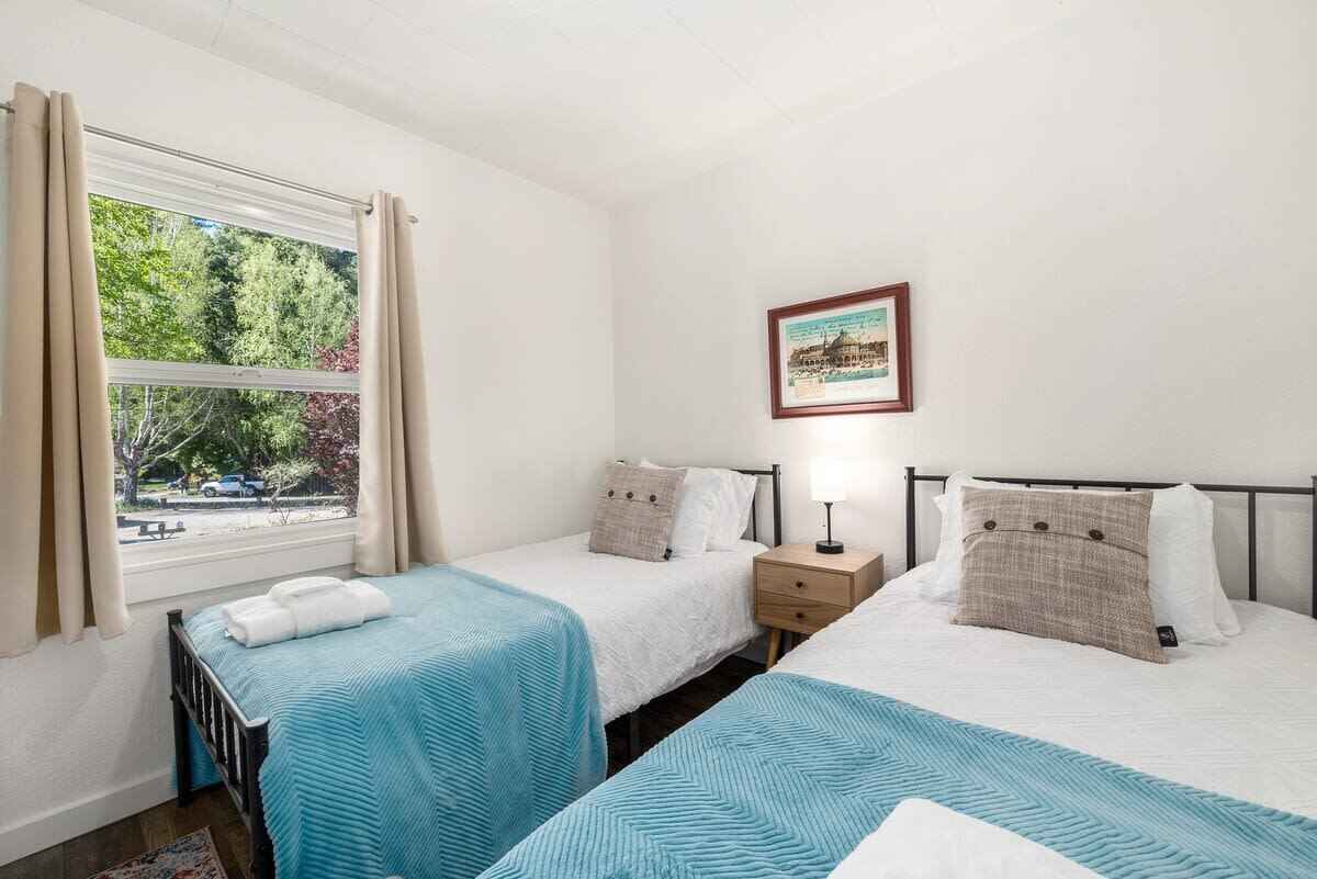 A hotel room with two single beds, a bedside table with a lamp, a framed picture on the wall, and a window with curtains.