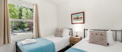 A hotel room with two single beds, a bedside table with a lamp, a framed picture on the wall, and a window with curtains.
