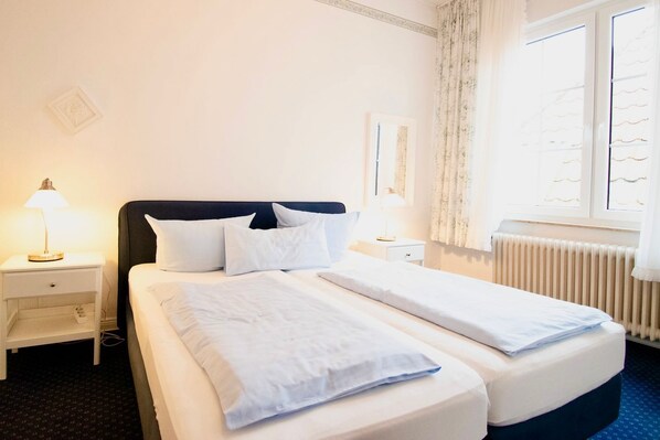 A hotel room with a double bed, two bedside tables, a nightstand, a window with curtains, and a radiator.