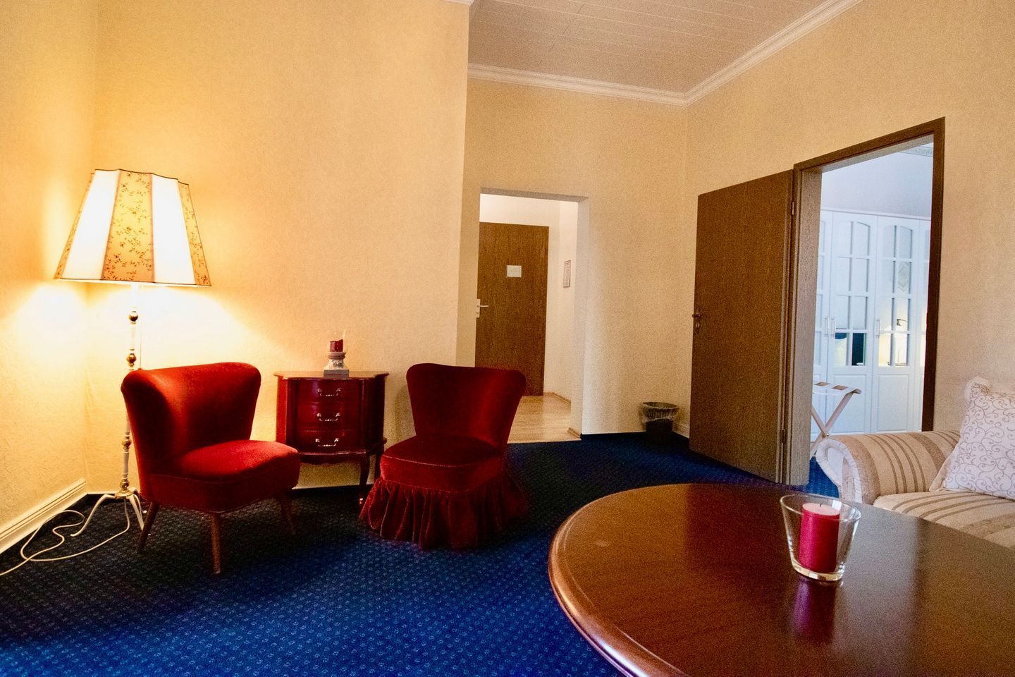 Ett hotellrum med ett blått heltäckningsmatta, två röda fåtöljer, ett träbord, en lampa, ett runt bord med ett glas på och en dörr som leder till ett annat rum.