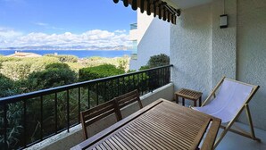 Ein Balkon mit Holztisch und -stühlen, einem kleinen Tisch und einer Liege. Der Balkon bietet einen schönen Blick auf das Meer und die entfernten Hügel.