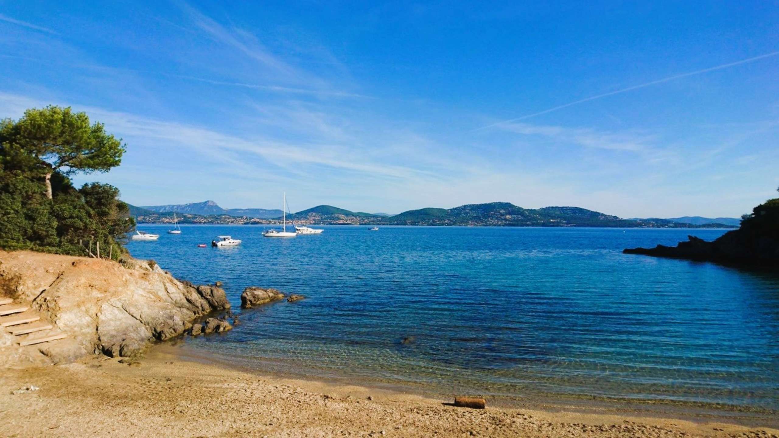 Une plage côtière avec des bateaux, une eau claire, un rivage rocheux et des collines à l’horizon.