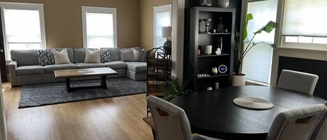 A modern living room with a sectional sofa, a dining table with chairs, a coffee table, a bookshelf, and a large window with blinds.
