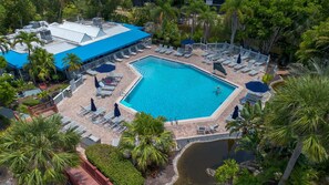 An aerial view of a resort with a large rectangular swimming pool surrounded by lounge chairs and umbrellas, with palm trees and a building with a blue roof in the background.