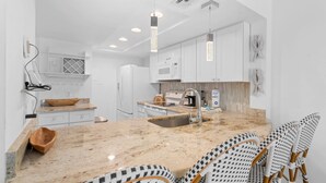 A modern kitchen with white cabinets, a granite countertop, and a built-in oven. There are two bar stools with checkered cushions. - First Floor Corner Condo at Park Shore Resort 149 (Naples)