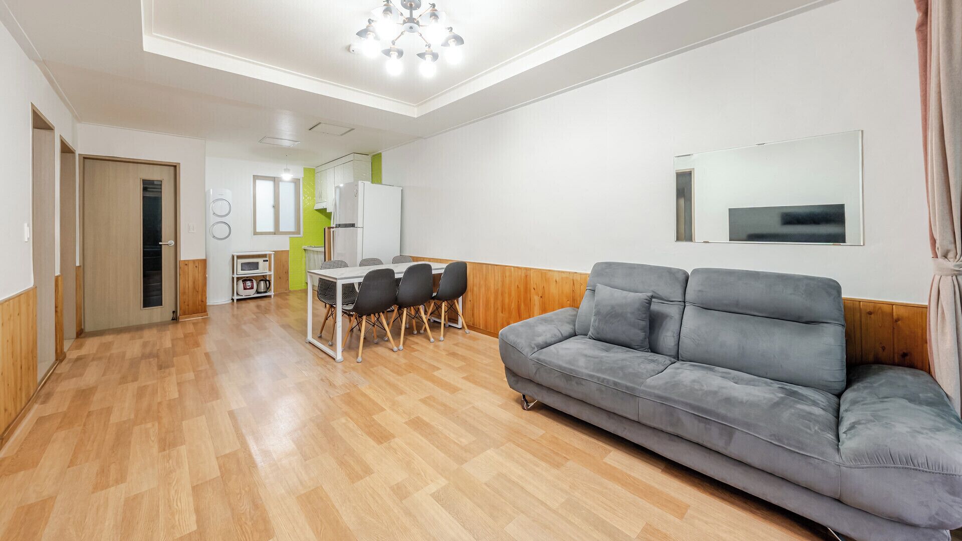 Una sala moderna con un sofá gris, piso de madera, un comedor con sillas, una cocina con microondas y un espejo grande en la pared.