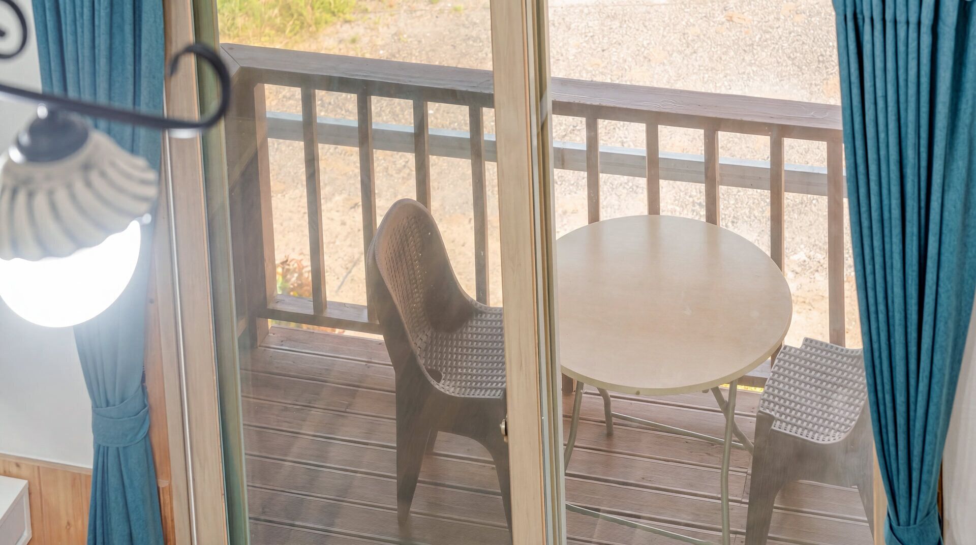 A wooden deck with a table and chairs, a view of a gravel area, and a curtain partially visible.