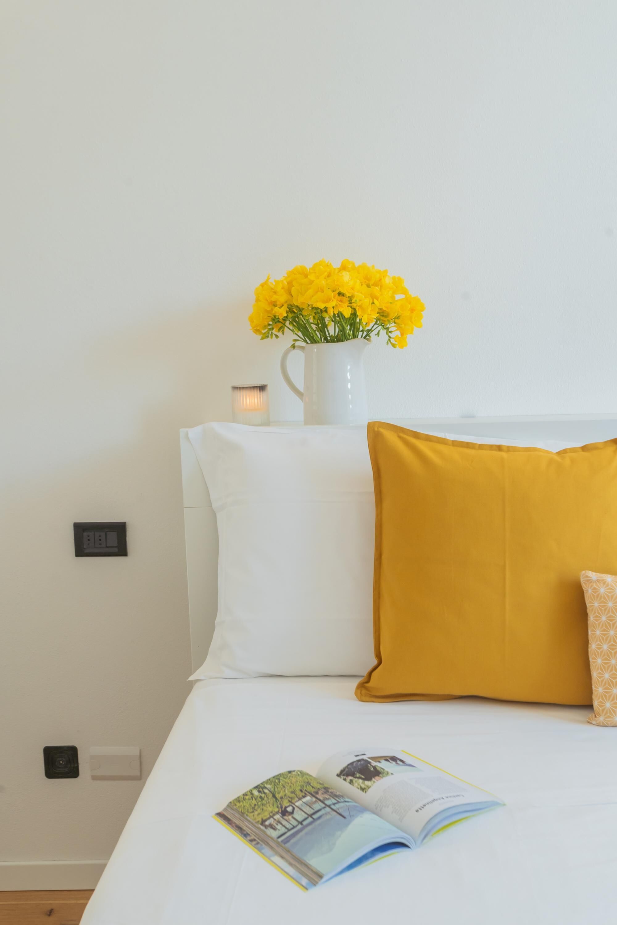 A neatly made bed with a white pillow, a mustard-colored pillow, and a small vase with yellow flowers on the headboard.