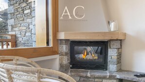 Una chimenea de piedra con leña encendida, un sillón de mimbre y una ventana con vistas a las montañas.