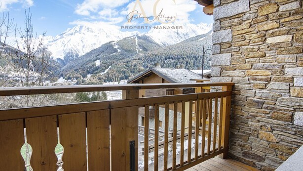 Un balcón de madera con pared de piedra, con vistas a un paisaje montañoso con cumbres nevadas.