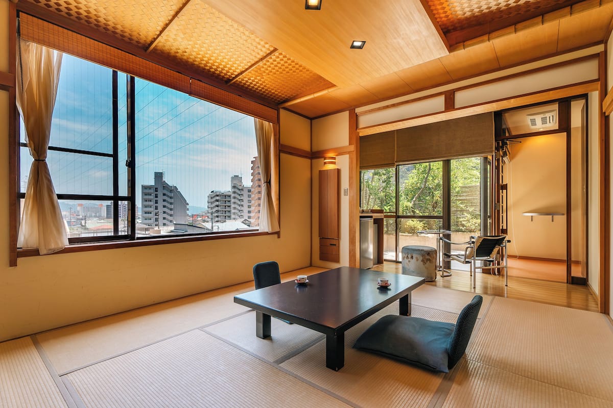 A traditional Japanese-style room with tatami flooring, a low table, and a view of the city through large windows.