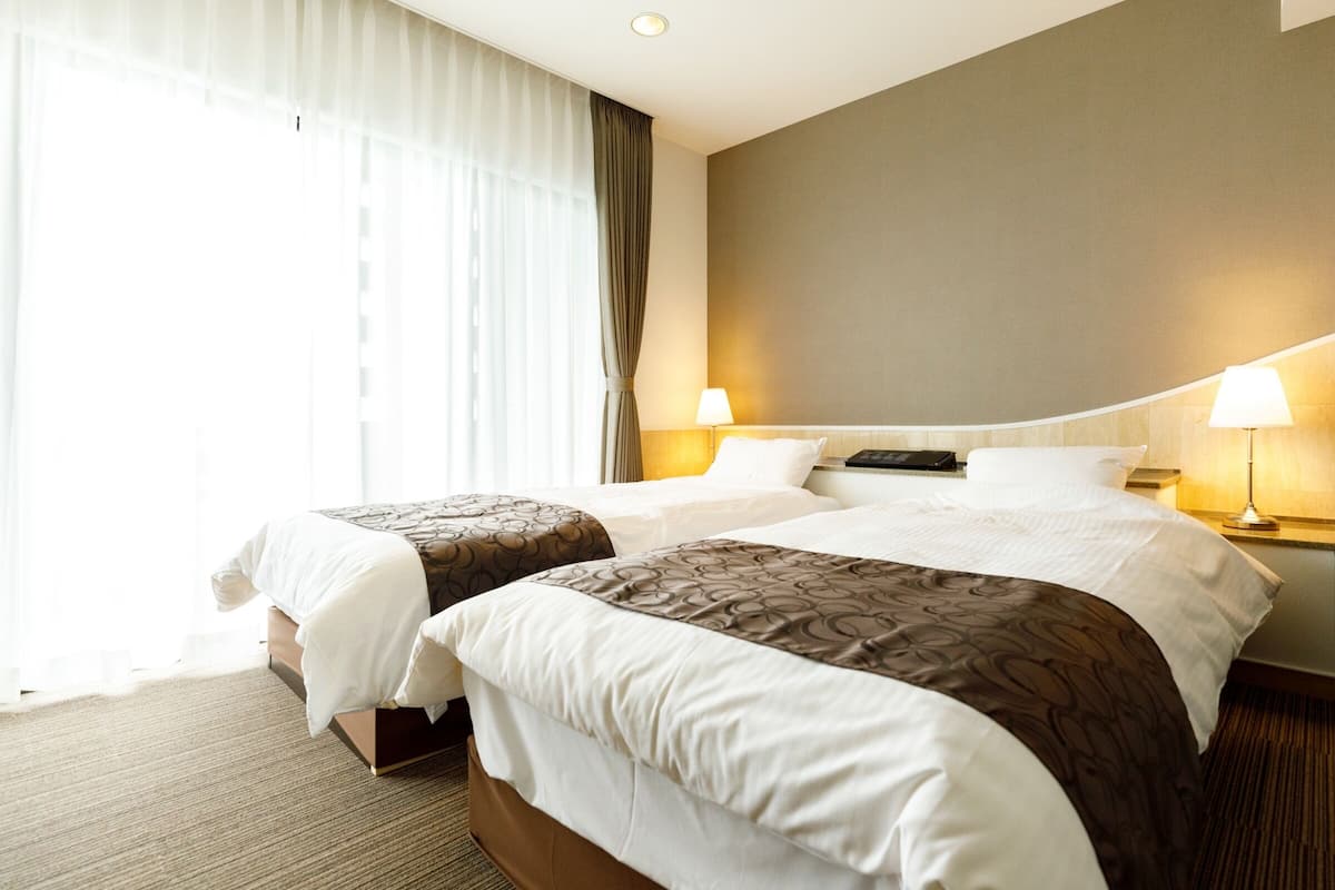 A hotel room with two beds, a large window with sheer curtains, and a wall-mounted lamp.