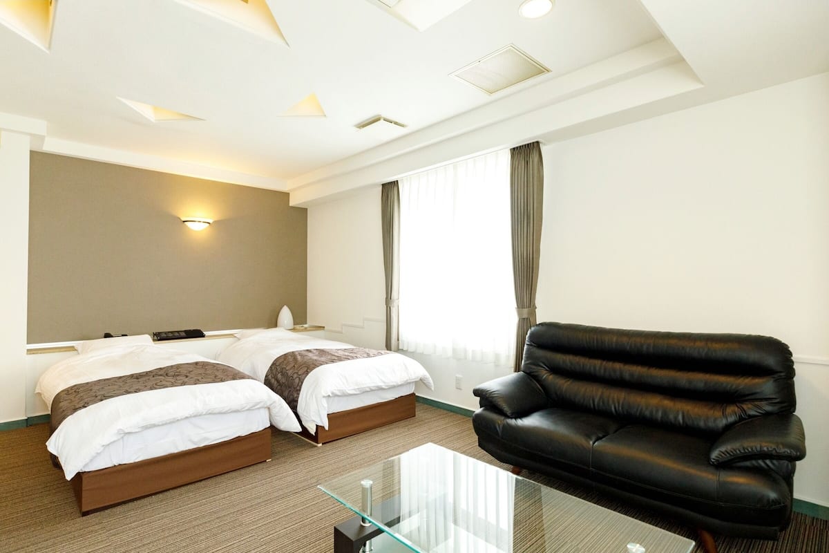 A hotel room with two beds, a leather sofa, a glass coffee table, and a large window with curtains.