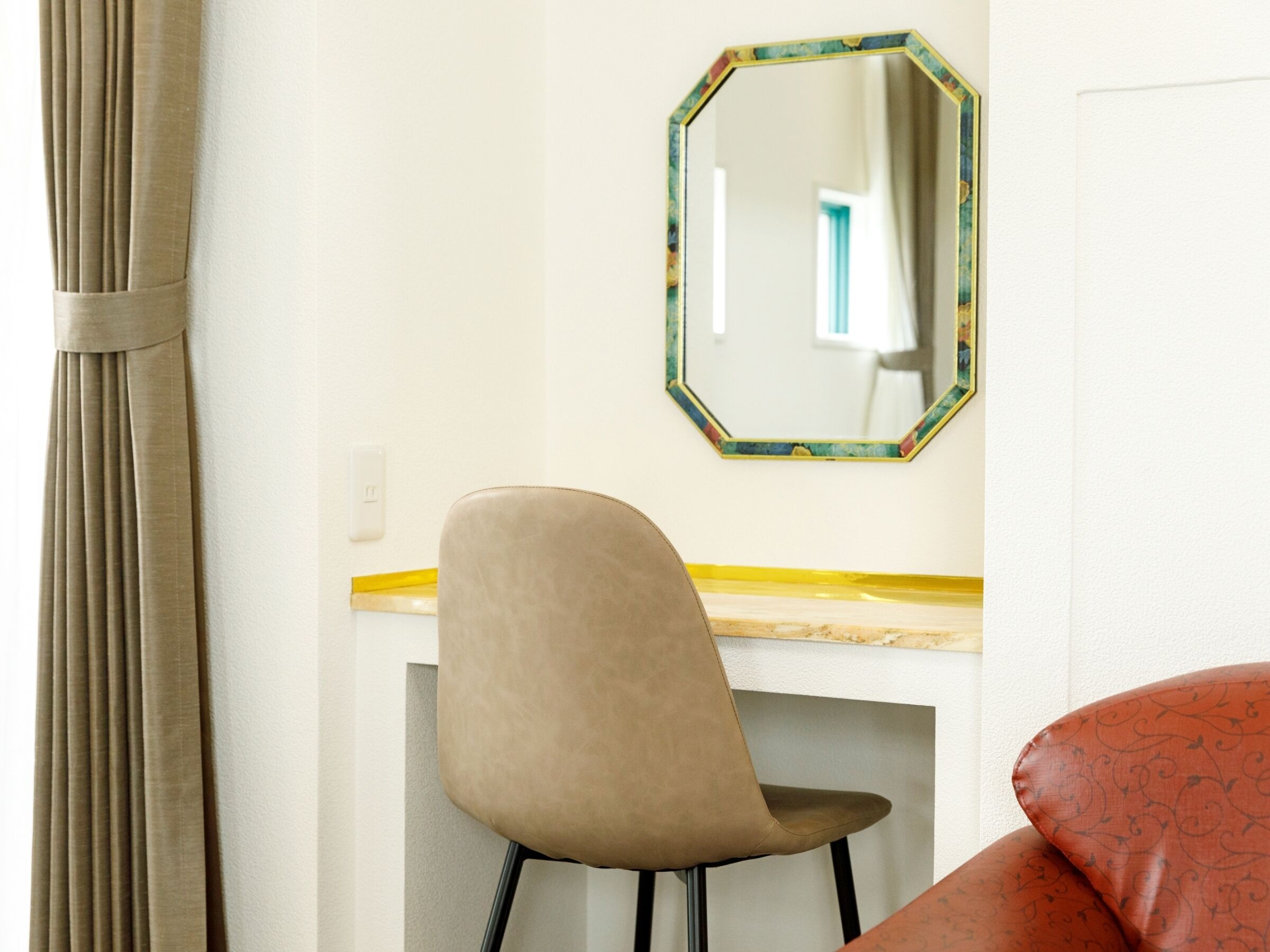 Quarto com uma cadeira bege, um espelho com moldura colorida e um balcão de madeira.