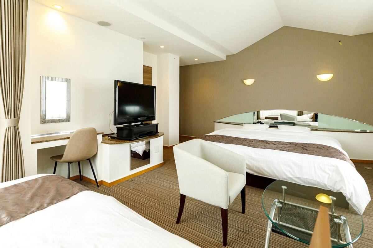 A hotel room with a bed, a desk with a television, a chair, and a glass table with a lamp.